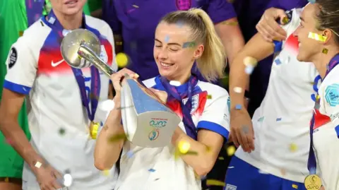 Reuters A woman smiles and looks at a silver trophy she is holding with both hands. She has blonde hair tied in a pony tail and is wearing a white England football shirt with red and blue detailing. the trophy says 'Euro' on the base. There is confetti in the air and other female English footballers in the background smiling and celebrating. They are all wearing gold medals tied to a purple ribbon around their necks.