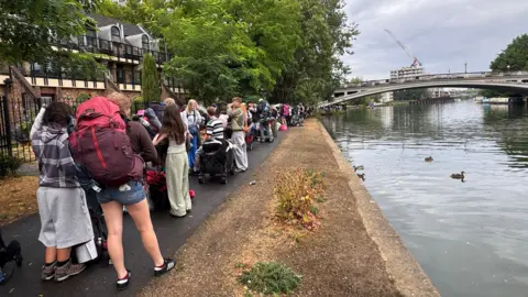 The River Thames is pictured on the right and a long line of people queueing for a boat carrying bags is pictured on the left. The weather is overcast.