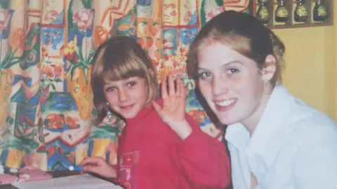 Supplied A girl and a young woman sitting at a table. The girl, to the left, has short blonde hair. She is wearing a bright pink jumper and waving at the camera. She is holding a pen and has a notebook in front of her. The woman has blond hair, tied back. She is wearing a white shirt. Behind them are blue, red, yellow and orange patterned curtains. 