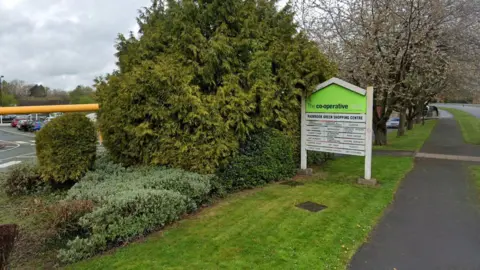 A Co-op food sign near a pavement. A car park can be seen in the background. There is a line of trees. 