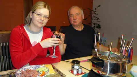 Handout A young woman in a red top clinks a glass with an older man in a black t-shirt while sitting at a table laid with a fondue set. She has a plate of food in front of her. 