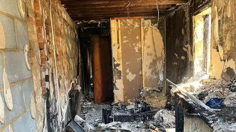 Inside of a flat destroyed by fire. Walls are bare concrete and brick with fire ash and debris across the room.