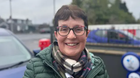 Karen McKenna - a woman with short brown hair smiles at the camera. She has a  pair of colourful square framed glasses on, as well as a plaid scarf and a green puffer coat on. She is standing in front of two parked blue cars and the background is blurred.