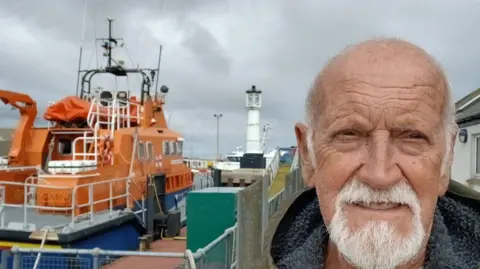 Derek Adams Derek Adams with short white hair and beard standing next to an orange lifeboat moored close to a white lighthouse on a black base.