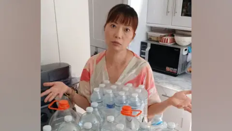 Pui Man is wearing a pink and white V-neck top. She has her hands raised by her sides - as if she is shrugging - and is pictured in her kitchen looking fed-up next to a pile of water bottles.
