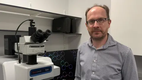 The photo shows a man in a black and white patterned shirt smiling at the camera. He's wearing glasses and has brown hair. He's standing in a laboratory, and is next to a microscope. Behind him is a computer screen, which has multi-coloured bubbles on it. 