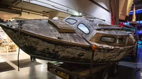 Clare James Photography The Hurley Burley yacht which is tilted at a slight angle in the middle of the museum. It is very dirty and brown and the words on the side of the boat have faded. It is resting on the back of a trailer.