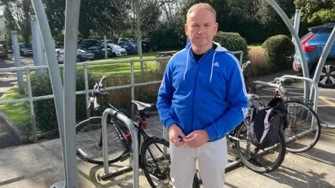 Councillor John Stephens stands in front of a bicycle rack on a sunny day in a car park which is full of vehicles. He is wearing a blue Adidas hooded jumper and white jeans.