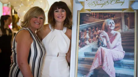 Angela MacVicar Smiling Angela MacVicar and Lindsay MacCallum wearing enactment      dresses posing adjacent  to a photograph   of Johanna MacVicar with helium-filled balloons down  them