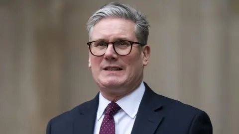 PA Media A man with grey hair and black-rimmed glasses photographed from the shoulders up. He is wearing a dark suit, white shirt and purple tie. 