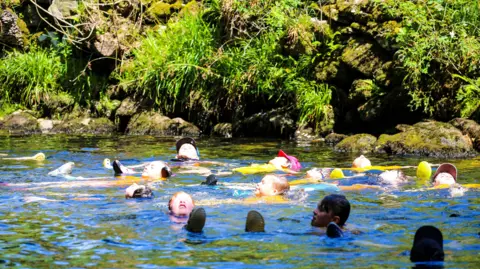 Grasmere Primary School A group of children floating in the water. They are wearing yellow polo shirts, with some wearing baseball caps and shoes. There are rocks and weeds on the riverbank.