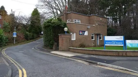 Geograph/JThomas The North entrance to Scarborough Hospital, with a building reading "Scarborough Hospital", behind a blue and white sign with "Scarborough Hospital" written on it and an NHS logo.