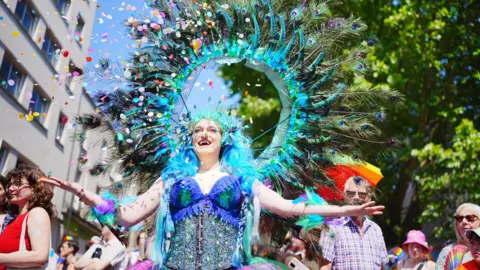 PA A person wearing a ring of blue peacock feathers on their back and a blue sequinned corset with a blue wig. They have their arms outstretched and confetti is falling down in front of them.