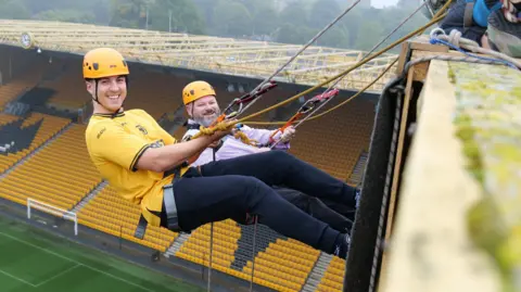 Two men wearing bright yellow helmets hold onto abseiling ropes above a football stadium. They are smiling at the camera.