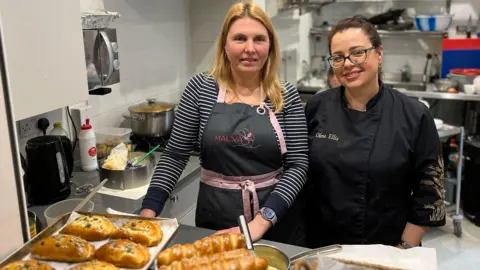 Iryna is blonde and is wearing a striped black and white top with a black apron with pink lining. Olena is wearing a black top with black glasses and brown hair. They are standing over the metal counter with baked goods on the counter.