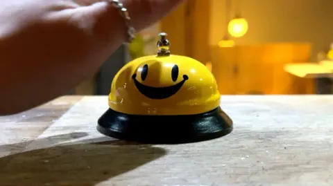 A hand is about to hit a bright yellow bell. It was a smiley face on it. There is a ring on the thumb.