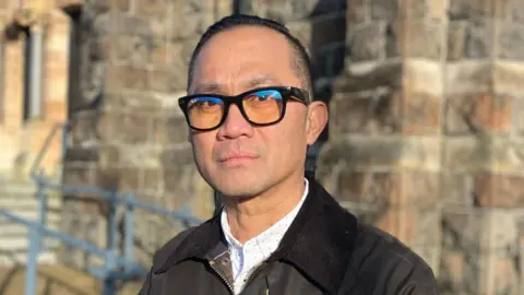 Aldrin Banas is standing outside a light stone chapel. He is wearing a black coat with a white shirt patterned with black polka dots. He has short black hair and is wearing heavy dark framed glasses. 