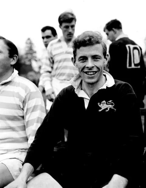 Getty Images A black and white photograph of Arthur Smith as a young man. He is smiling broadly and wearing a dark-coloured rugby top. he is surrounded by others in similar clothes. 