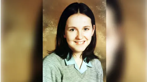 McFarland Family Samantha McFarland smiles at the camera with should length dark hair, green eyes and is wearing a grey sweater with a blue shirt beneath.
