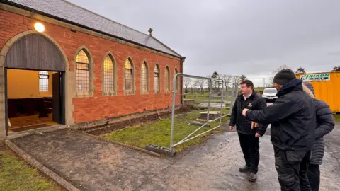 Angled side view of the chapel at Dudley Cemetery - a low brick building with a slate roof and arched windows. A temporary fence surrounds it, with three people viewing the structure. Part of a temporary site cabin can be seen behind them.