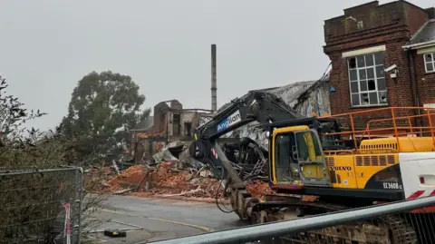 Demolition plant at the site of the former factory. Part of the roof has collapsed and the ruins can be seen in the background 