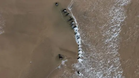 David McGloin Aerial shot of the wooden structure on the beach. A wave passes over a number of wooden pieces that look to be in the shape of a section of a boat.