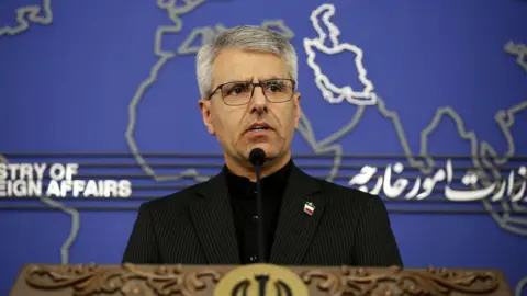 A man in black suit jacket speaks at a podium with a blue background 
