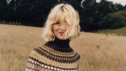 Maisie Peters wears a high neck brown and yellow knit jumper and has her blonde hair cut into a wavy bob. She stands against a field with crops and sheep