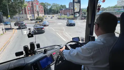 A photograph looking over a coach driver’s shoulder driving along a road in Stroud. The driver is wearing a light-coloured shirt and glasses. He is holding the steering wheel with the dashboard, controls and mounted devices visible. Through the windscreen, several cars, road markings, street signs and a red-brick building can be seen, with trees and lampposts lining the road in bright daylight.
