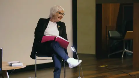 Think Human Actress Annette Badland on stage holding a red book. She appears to be laughing as she looks at something out of frame. There is a wooden table with some books on it. There is a small light in front of her.