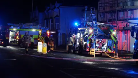 Two fire engines with blue lights in parked on the road where the fire broke out. Fireman stand beside the vehicles getting equipment and hoses.