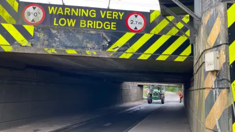 A green tractor in the distance has a badly damaged roof and wing mirrors. It is on a road under a bridge and there is debris and shattered glass on the road and pavement. There is sign a on the bridge that reads "warning very low bridge".