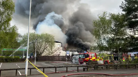 Fire engines in a car park with thick black smoke rising above a white sided building