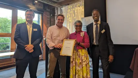 Handout Mr Brown stands smiling in a line with his colleagues - two men and a woman. A man and a woman in the middle of the line are holding a gold framed certificate. 