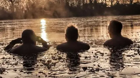 Kellie Tarr Three people submerged up to their shoulders in icy water, during sunrise.