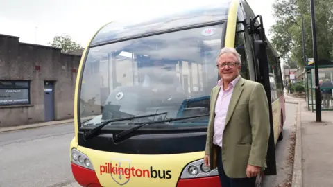 LCC County councillor Rupert Swarbrick standing smiling at a bus stop in front of a yellow and red bus