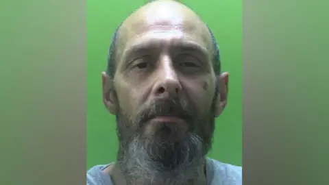 Mugshot of Jason Burton, a Caucasian, balding, 48-year-old man with a greying beard and a bruise on his left cheek.