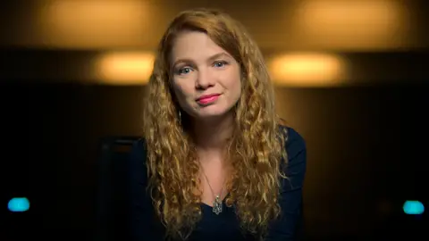 Alastair McKevitt A woman sits against a dimly lit amber background, in an interview-type set up. She's smiling slightly, her long, curly red hair cascading down her shoulders.