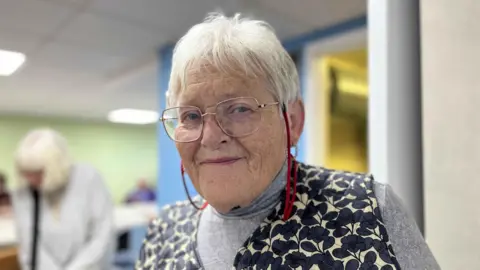 George Carden/BBC Chris Elshabba inside a brightly lit room wearing a floral top and glasses. she has short grey hair