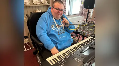 Stephen Farlow is wearing a blue hoody and sits at his keyboard. He has glasses and short brown, greying hair. He holds a mic in his hand. 