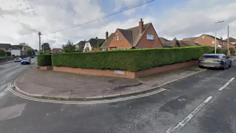 Google Maps Entrance to Briar Close, Exmouth. There is a main road to the left with a smaller road with a large hedge at the entrance. A small brick wall has a sign that reads "Briar Close" on it.