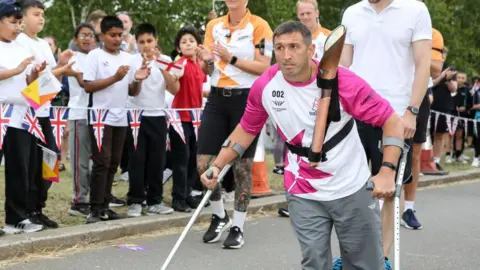 Nick England/Getty A man is walking with crutches, with a torch strapped around his chest. Crowds are cheering him on. He is wearing grey trousers and a white and pink top.