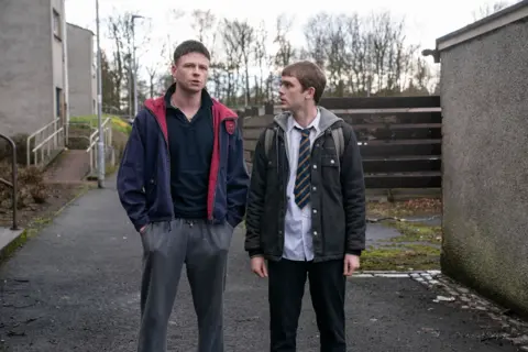 BBC/Mam Tor Productions/Anne Binckebanck Outside an estate, a teenager in a polo shirt, tracksuit bottoms and a blue jacket with red lining looks forward as a shorter teenager in a school uniform looks at him.