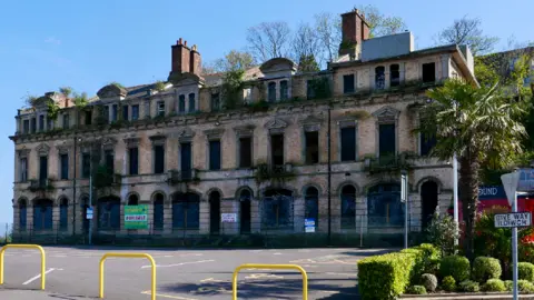 Morgan Ellis Leah Image of the Marine Hotel taken from further back. It is a large light brown brick building with greenery growing out of many of the windows. On the fence surrounding the building is a green 'for sale' sign.
