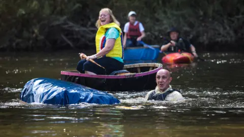 One man with balding hair is pictured in the water next to a capsized coracle. A woman in a yellow life jacket remains on her craft behind. A third coracle and rider is in the background, alongside a man in a kayak.