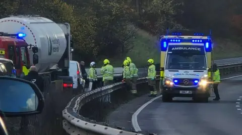 An ambulance with the lights on stopped in the middle of the road. Staff members are seen around it. There is a lorry behind the guardrail.