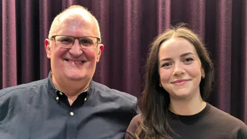 Spencer and Hollie stand side by side in front of a purple curtain, smiling to the camera. Spencer is a middle-aged, balding man with glasses, wearing a dark blue shirt. Hollie is a young woman with long brown hair, wearing a brown top.