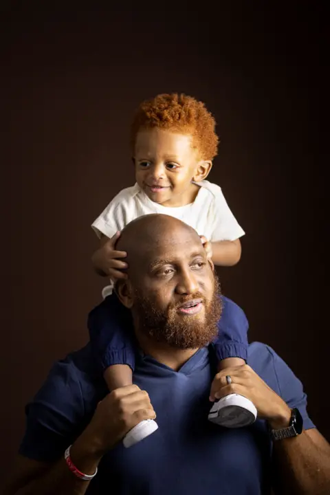 Kieran Dodds Studio portrait of an adult wearing a blue shirt with a young child seated on the adult’s shoulders. The child wears a light-colored T-shirt, dark trousers, and white shoes, with hands resting on the adult’s head. Both are photographed against a dark brown background with warm, soft lighting.
