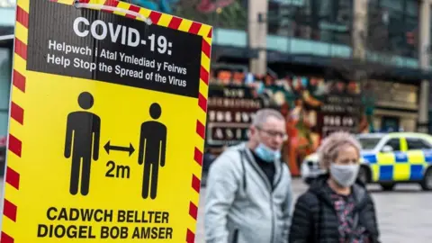 Getty Photo of two people walking in Cardiff next to a social distancing sign. A police car can be seen behind them.