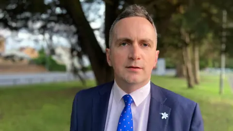 A man in a navy suit, with short grey hair, pictured against a tree